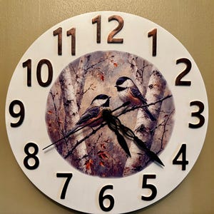 May include: Round wall clock with a white face and black numbers. The center features a painting of two birds perched on a branch in a forest setting. The clock hands are black.