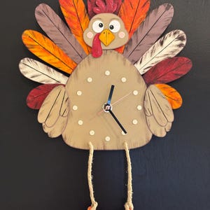 May include: A turkey-shaped wall clock with a tan body, white dot hour markers, and black clock hands. The turkey features a colorful fan of feathers in red, orange, brown, and white, with dangling legs made of tan rope and orange feet.