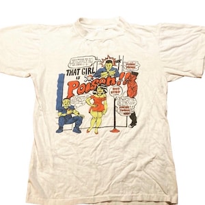 Op de afbeelding: Een wit t-shirt met korte mouwen en een kleurrijke grafische print in stripstijl. Het ontwerp bevat drie figuren, een bushaltebord en de tekst "That Girl is Poison!!" samen met diverse tekstballonnen met dialoog.