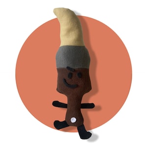 May include: A soft toy paintbrush with a beige bristle, grey ferrule, and brown handle. The toy has a smiling face, black arms and legs, and is set against an orange circle background.