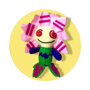 Peut inclure: Poupée fleur en peluche faite à la main avec un design fantaisiste. La poupée a un visage crème avec des yeux en spirale rouges et une bouche souriante. Les pétales de la fleur sont roses avec des rayures magenta. Le corps de la poupée est vert et bleu.