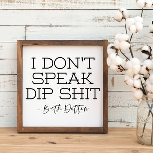 May include: A white wooden sign with a brown frame, featuring the text "I don't speak dip shit" and the signature "-- Beth Dutton". The sign is displayed on a wooden surface with a vase of white cotton flowers in the background.
