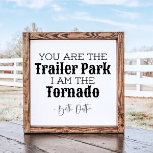 May include: A framed wooden sign with the quote "YOU ARE THE Trailer Park I AM THE Tornado - Beth Dutton" in black text on a white background. The frame is a dark brown color, and the sign is displayed outdoors.