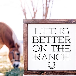 May include: A wooden sign with the text "Life is better on the ranch" and a horseshoe graphic. The sign is in front of a blurry image of a brown horse.