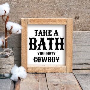 Peut inclure: Un panneau en bois rustique avec le texte "TAKE A BATH YOU DIRTY COWBOY" en lettres noires. Un bocal en verre envelopp&eacute; de ficelle contient des tiges de coton. Le fond est en bois vieilli.