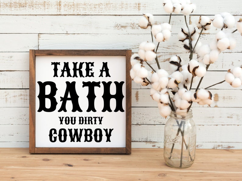 Peut inclure: Un panneau en bois encadr&eacute; avec le texte "TAKE A BATH YOU DIRTY COWBOY" en lettres noires. Un bocal en verre avec des tiges de coton est &agrave; droite du panneau. L'arri&egrave;re-plan est un mur en bois blanc.