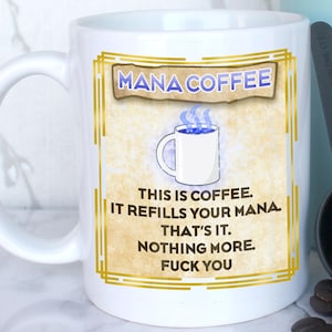 May include: A white coffee mug with the text "MANA COFFEE" and a humorous message. A light blue French press and a black scoop with coffee beans are next to the mug. The mug has a gold border.