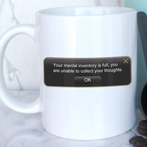 May include: White coffee mug with a black and gold pop-up message that reads "Your mental inventory is full, you are unable to collect your thoughts. OK" with a red X in the top right corner.  A blue French press and a black scoop with coffee beans are in the background.