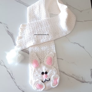 May include: A white crocheted scarf featuring a bunny face with pink inner ears and paws, and black button eyes. The scarf has a fluffy white tail and is made of a soft, textured yarn. The scarf is designed for children.