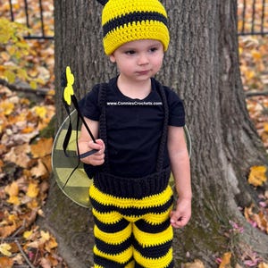 May include: A child's crochet bee costume, featuring a yellow and black striped hat with antennae, matching striped pants, and a black shirt with suspenders. The costume includes a yellow flower wand and bee wings.