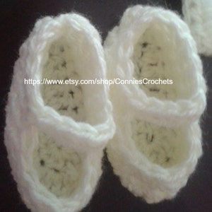 May include: A pair of white crocheted baby shoes with a strap across the top. The shoes are made with a delicate stitch and have a soft, comfortable feel.