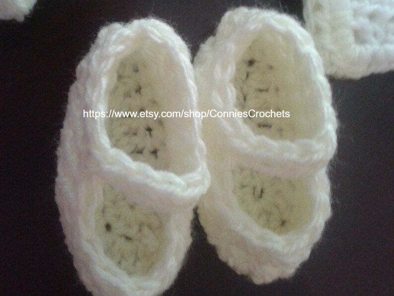 May include: A pair of white crocheted baby shoes with a strap across the top. The shoes are made with a delicate stitch and have a soft, comfortable feel.