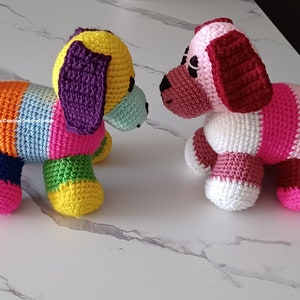 May include: Two crocheted stuffed dog toys. One dog is multi-colored with a purple ear and the other dog is pink and white with a red ear.