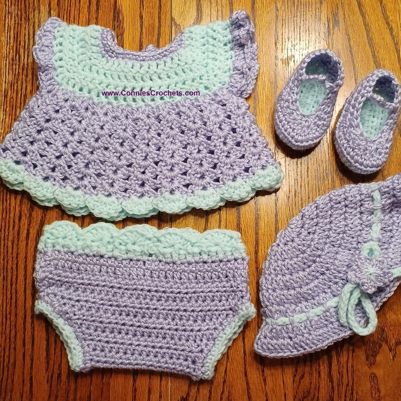 May include: A crocheted baby outfit set in lavender and mint green. The set includes a dress, diaper cover, hat, and shoes. The dress has a scalloped edge and the hat has a bow. The text "www.ConniesCrochets.com" is visible on the dress.