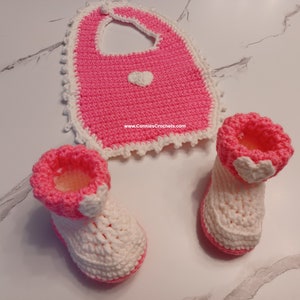 May include: A set of pink and white crocheted baby booties and a bib with a heart design. The bib has a scalloped edge and the booties have a white heart on the top.