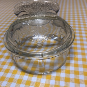 May include: A clear glass dish with a lid. The lid has a decorative handle. The dish is round and sits on a yellow and white checkered surface. The glass has a textured appearance.