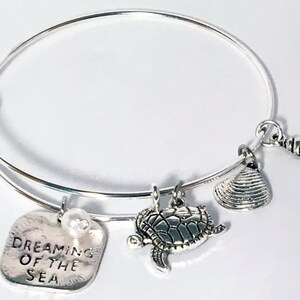 Può includere: Bracciale a cerchio color argento con ciondoli a tema marino. I ciondoli includono una tartaruga, una conchiglia e un ciondolo quadrato con la scritta "Dreaming of the Sea" e una piccola perla. Il bracciale è regolabile.