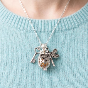 May include: A silver-toned necklace featuring a central cage pendant containing a small amber-colored sphere, accompanied by a heart-topped key charm and a detailed dragonfly charm, all suspended from a delicate silver-toned metal chain.