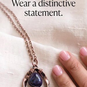 May include: A copper-toned necklace featuring a dark purple, teardrop-shaped stone set within a hammered, triangular metal frame. The text TRENDYGURL, Escape the mass-produced. Wear a distinctive statement. is visible at the top of the image.