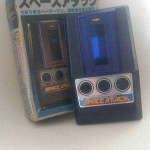 Puede incluir: Un juego electrónico de mano azul y negro llamado "Space Attack" de Tomy Electronics. El juego tiene una pantalla pequeña y tres botones. La caja del juego está en japonés y muestra una imagen del juego.