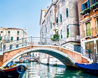 May include: A view of a canal in Venice, Italy. A small boat is in the foreground, and a bridge arches over the canal. The buildings on either side of the canal are old and have a weathered look.