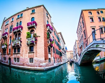 May include: A view of a narrow canal in Venice, Italy. The canal is lined with colorful buildings, some with flower boxes. A stone bridge arches over the canal.