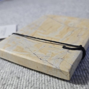 Peut inclure: Un dessous de verre rectangulaire en pierre de couleur claire, entouré d'un cordon noir. Le dessous de verre a un aspect marbré avec de subtiles variations de couleur et de texture. Une petite étiquette est attachée.