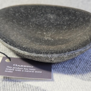 Peut inclure: Un plat en pierre de gabbro ovale de couleur gris foncé. Le plat a une surface texturée et une étiquette attachée avec le mot "Gabbro" et des informations sur le produit. Le plat est scellé avec un scellant en pierre naturelle.