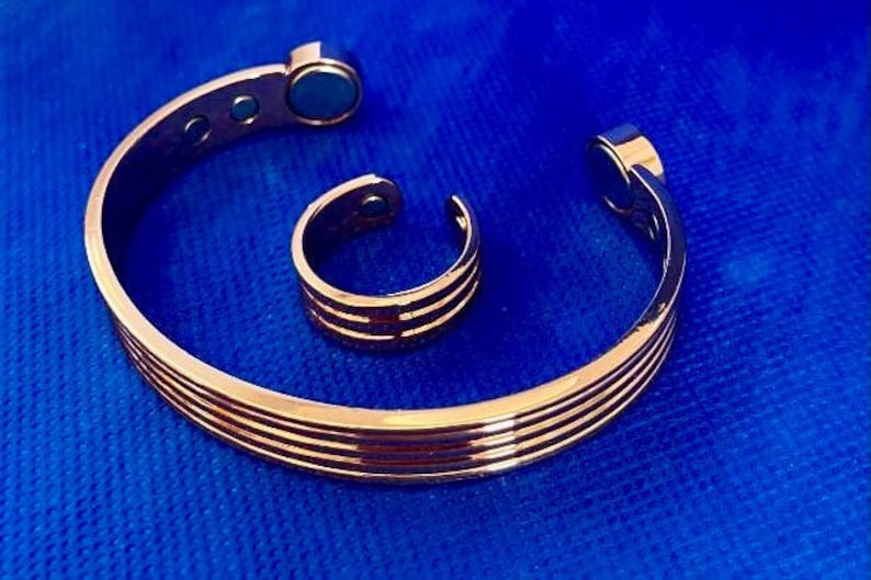 May include: A set of two copper-toned magnetic bracelets. The larger bracelet is a bangle style with a magnetic clasp. The smaller bracelet is a ring style with a magnetic closure. Both bracelets have a polished finish.