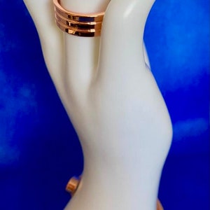 May include: Two gold-toned rings on a white mannequin hand. The top ring is a simple band, while the bottom ring is a wider band with a textured design.