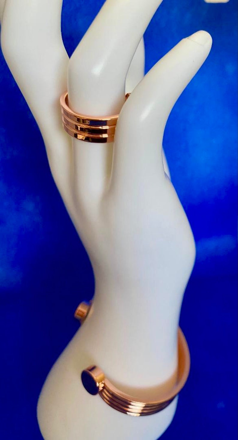 May include: Two gold-toned rings on a white mannequin hand. The top ring is a simple band, while the bottom ring is a wider band with a textured design.