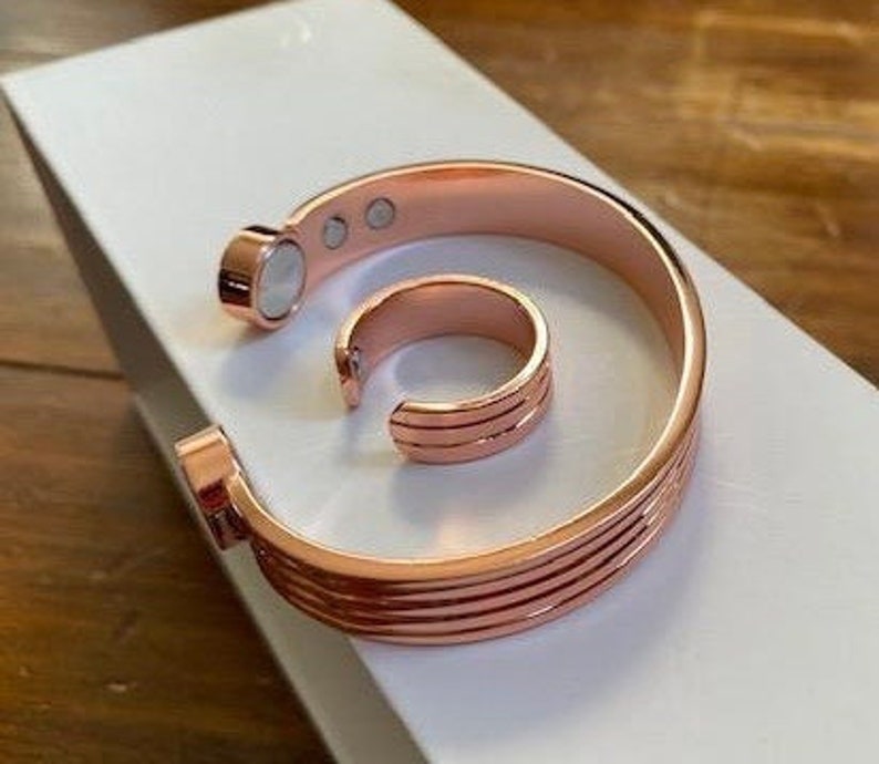 May include: A set of two copper magnetic bracelets. The bracelet is a wide band with multiple ridges. The ring is a simple band with a smooth surface. Both pieces are rose gold in color.