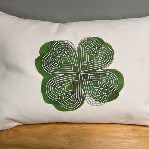 May include: White pillow with a green four-leaf clover design. The clover is outlined in white and has a Celtic knot pattern.