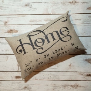 May include: A beige pillow with the word "Home" printed in black script. The pillow also has the coordinates "35° 9' 28.1304, N 106° 29' 15.3024''" printed in black.