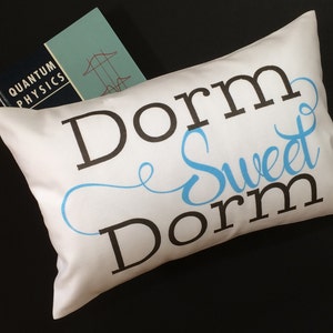 May include: White pillow with the text "Dorm Sweet Dorm" in black and blue lettering. The pillow is sitting on a black surface with a book titled "Quantum Physics" on top of it.
