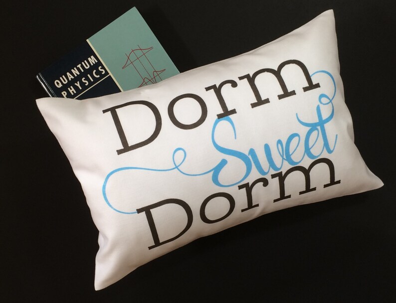 May include: White pillow with the text "Dorm Sweet Dorm" in black and blue lettering. The pillow is sitting on a black surface with a book titled "Quantum Physics" on top of it.