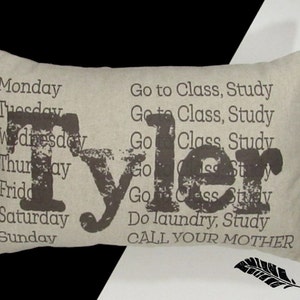 May include: Rectangular beige pillow with the name "Tyler" in large, distressed brown letters. The days of the week and daily activities are printed in a smaller font. A black feather graphic is in the lower right corner.