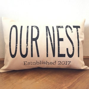 May include: A white throw pillow with black text that reads "OUR NEST Established 2017".