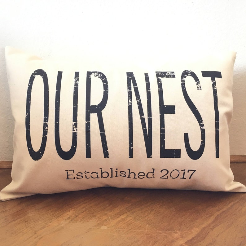 May include: A white throw pillow with black text that reads "OUR NEST Established 2017".