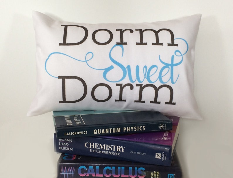 May include: White pillow with the text "Dorm Sweet Dorm" in black and blue lettering. The pillow is sitting on top of a stack of textbooks.