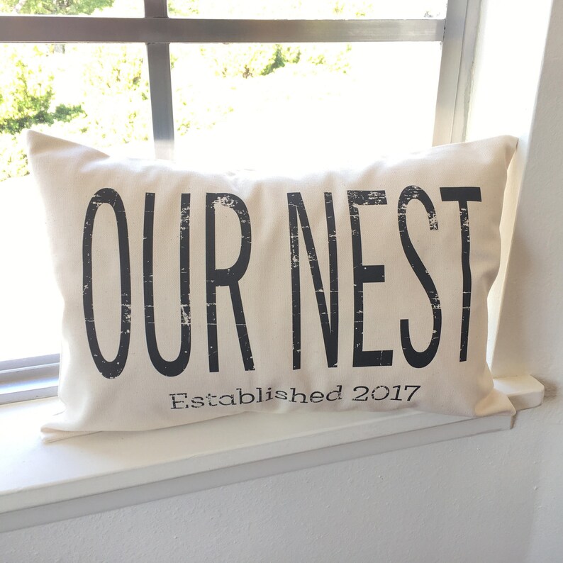 May include: A white throw pillow with black text that reads "OUR NEST Established 2017".