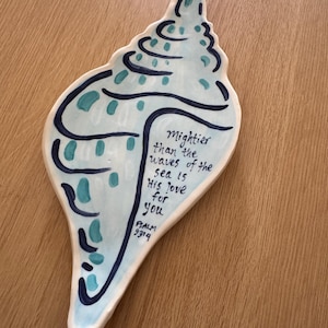 May include: A seashell-shaped ceramic dish with a light blue base and dark blue and teal accents. The dish features the text "Mightier than the waves of the sea is His love for you" and "Psalm 93:4".