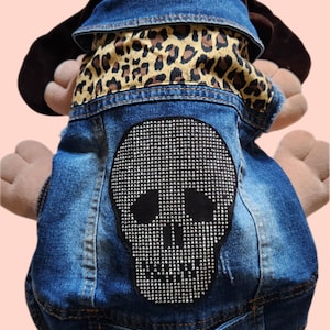 May include: A blue denim dog jacket featuring a leopard print collar and a rhinestone skull design on the back. The jacket has two button closures and a distressed appearance.