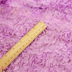 May include: Close-up of lavender fabric with a 3D floral rose design. A wooden ruler, marked in inches and centimeters, is placed diagonally across the fabric. The fabric has a delicate, textured appearance.