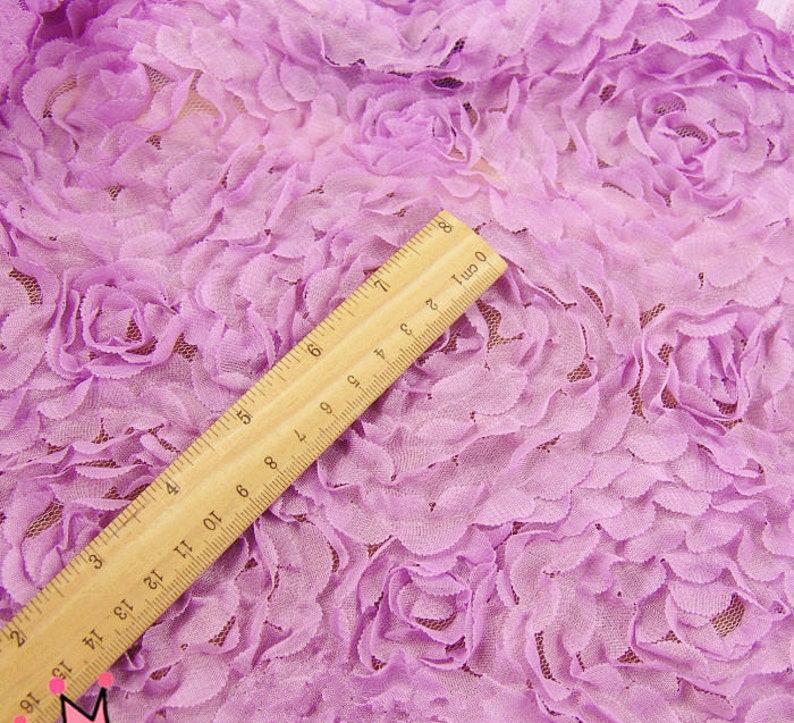 May include: Close-up of lavender fabric with a 3D floral rose design. A wooden ruler, marked in inches and centimeters, is placed diagonally across the fabric. The fabric has a delicate, textured appearance.