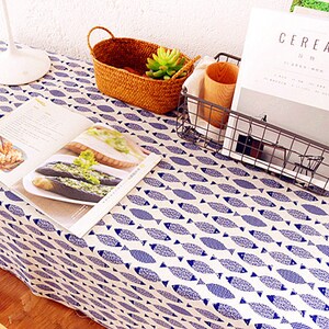 May include: A white tablecloth with a blue fish pattern. A cookbook, a woven basket with a small plant, and a wire basket with a wooden cup and a magazine are on the table.