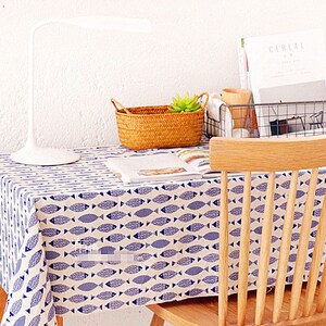 May include: A white table with a blue and white fish patterned tablecloth. A wooden chair with a slatted back is positioned at the table. A white lamp, a wicker basket with a green plant, and a wire basket with magazines are on the table.