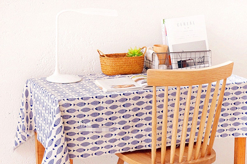 May include: A white table with a blue and white fish patterned tablecloth. A wooden chair with a slatted back is positioned at the table. A white lamp, a wicker basket with a green plant, and a wire basket with magazines are on the table.