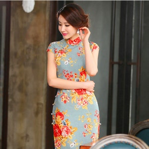 Puede incluir: Vestido azul claro de manga corta con cuello mandar&iacute;n y estampado floral. El vestido presenta flores rojas y amarillas con hojas verdes. El vestido tiene una silueta ajustada y detalles de botones en el lateral.