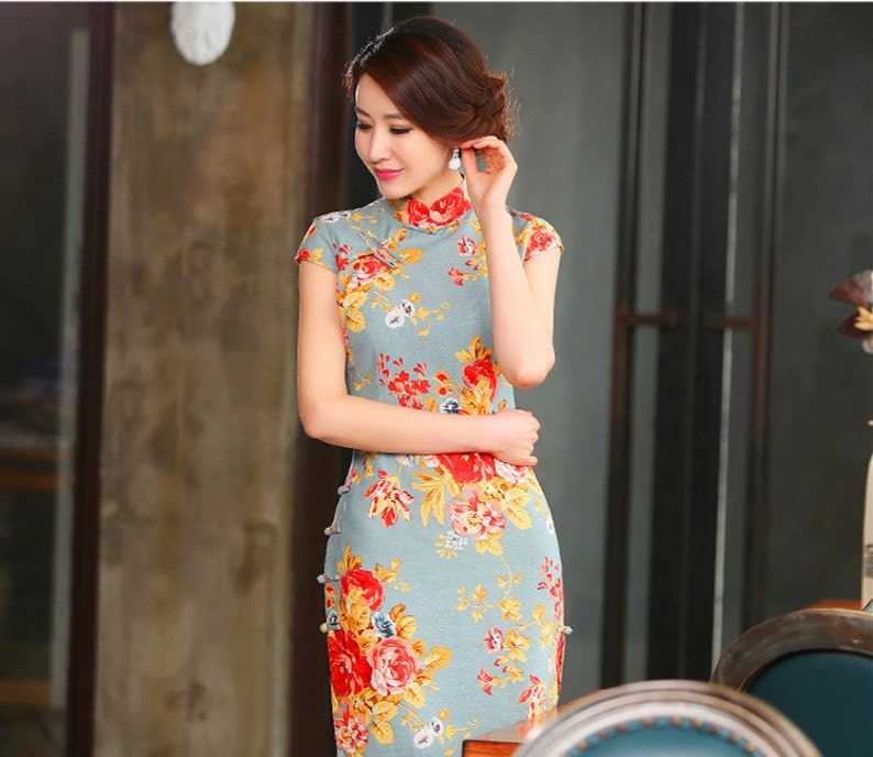 Puede incluir: Vestido azul claro de manga corta con cuello mandar&iacute;n y estampado floral. El vestido presenta flores rojas y amarillas con hojas verdes. El vestido tiene una silueta ajustada y detalles de botones en el lateral.